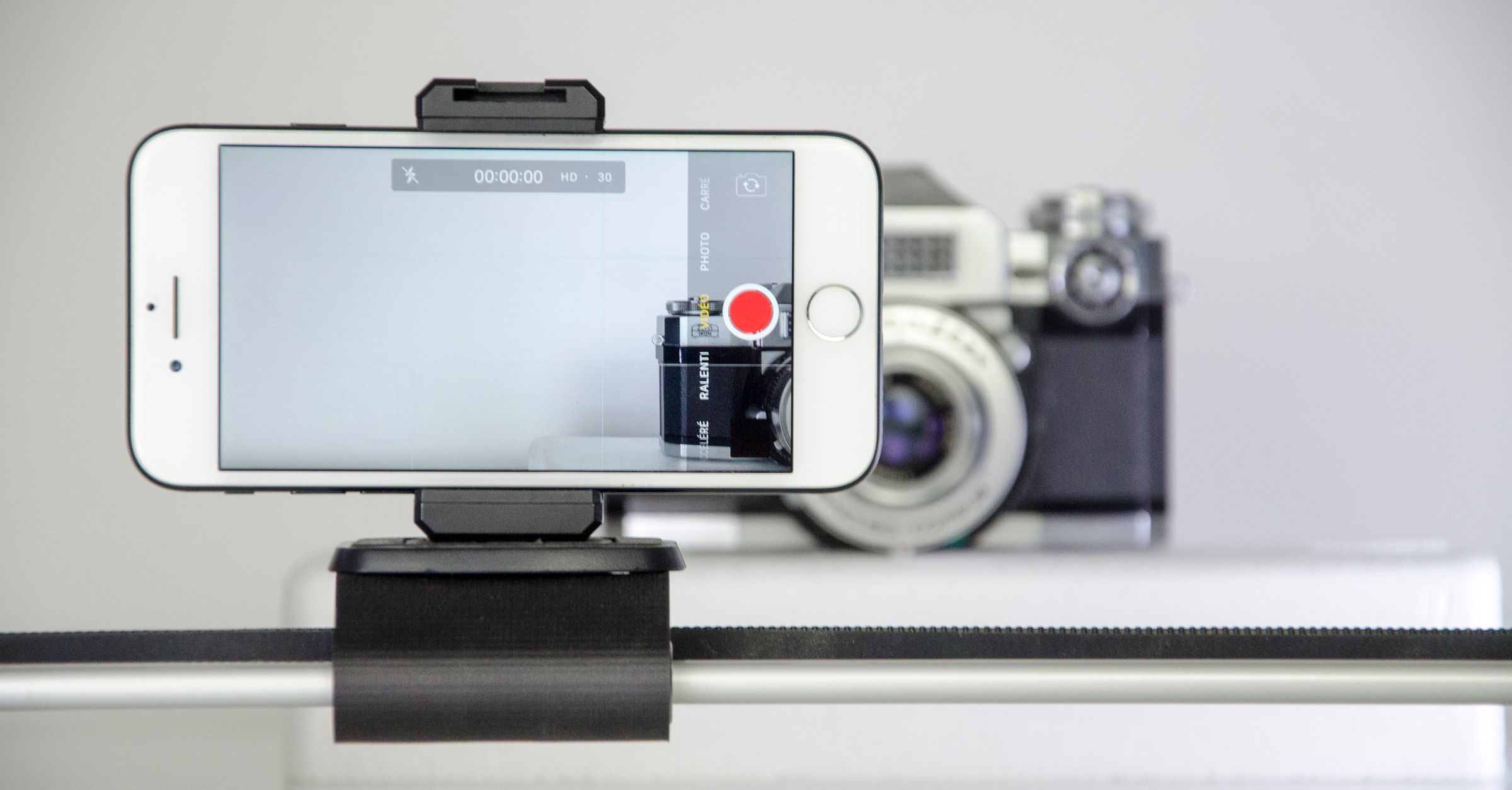 iPhone mounted horizontally in a clamp with a video recording interface, framing a camera in the background.