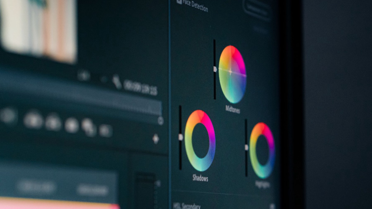A close-up of the color wheels in a video editing application on a dark monitor, labelled Shadows, Midtones and Highlights, with the timeline out of focus behind them.